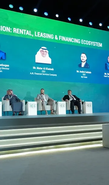 Five panelists seated on stage during a panel discussion with a large speaker backdrop screen in Riyadh
