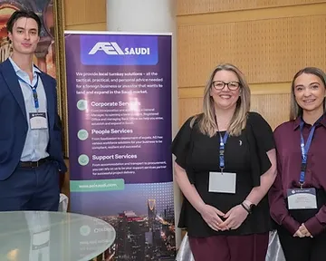 Three event delegates posing in front of a partner roll-up banner at a conference in Riyadh